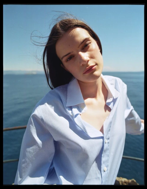 Portrait of a young woman in a striped shirt on a balcony overlooking the sea, captured in natural light on a sunny day.