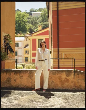 Model standing outdoors on a cobblestone terrace wearing a white long-sleeve shirt and white trousers in an Italian town street.