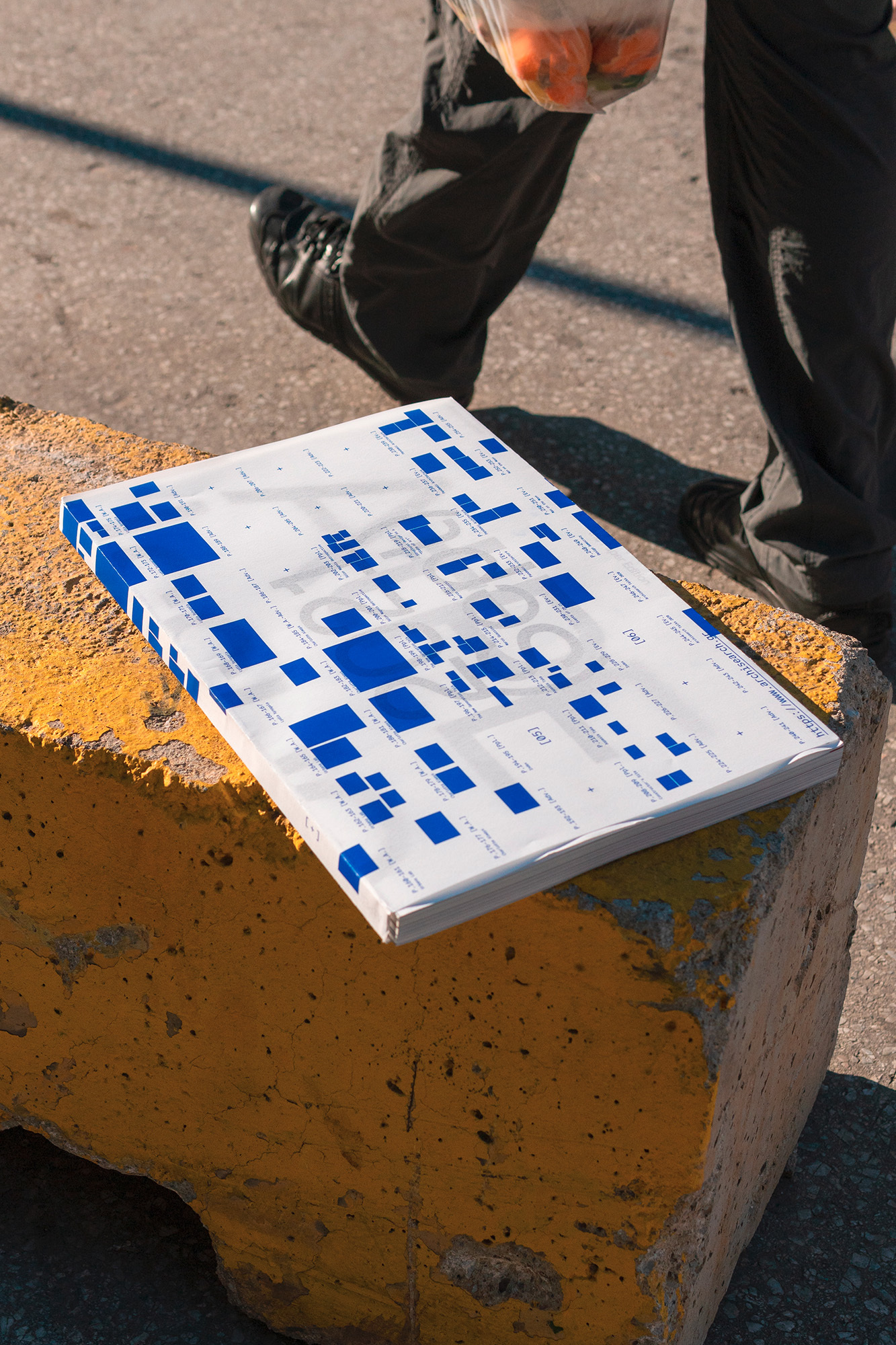 Book with a blue and white pixelated design sits on a yellow concrete block next to a walking person carrying a bag of oranges.