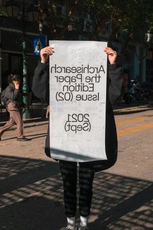 Person in black clothes holds a large newspaper with white background standing on a city street, hiding their face.