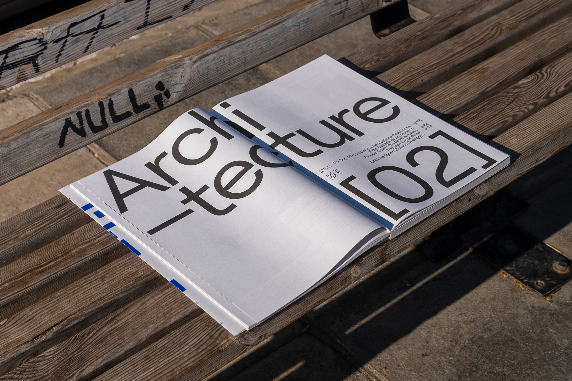 Open architecture magazine with bold black typography on a wooden park bench in bright daylight, showcasing the crisp details and textures.