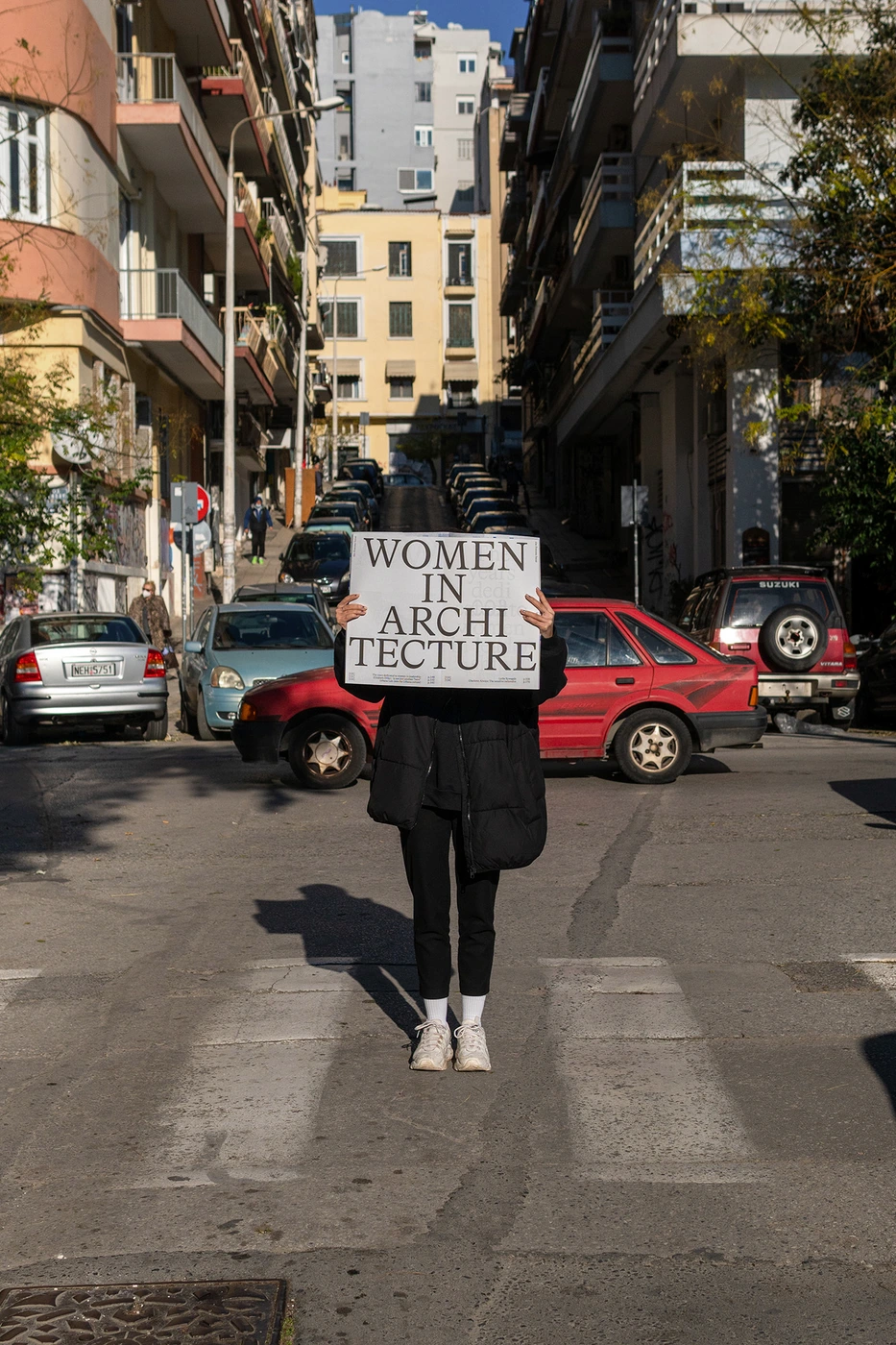 Person holding a sign reading Women in Architecture while standing on a crosswalk in a dense urban street.