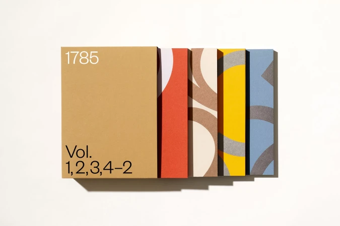 Five bound volumes with graphic patterns, in orange, brown, yellow, and blue, sit on a white surface in horizontal composition.