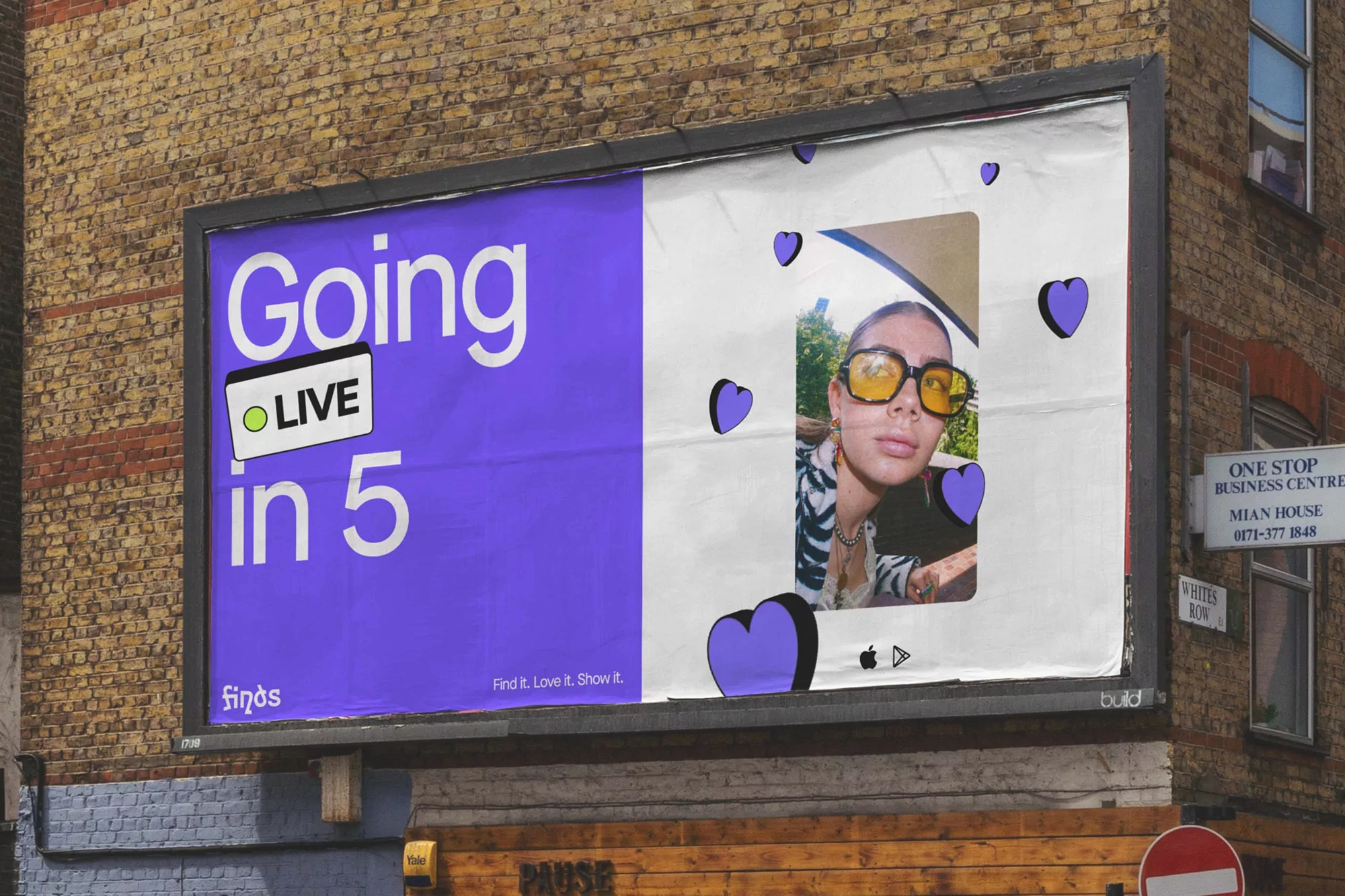 Finds billboard on a brick building with the text “Going Live in 5” and a woman with yellow glasses surrounded by purple hearts.
