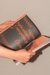Hands holding an open magazine displaying typography with the text Secret Signs Subculture Conspiracy Class against a plain neutral background