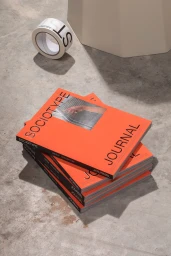 A stack of bright orange Sociotype Journal magazines on a concrete floor with branded tape and a geometric prop.