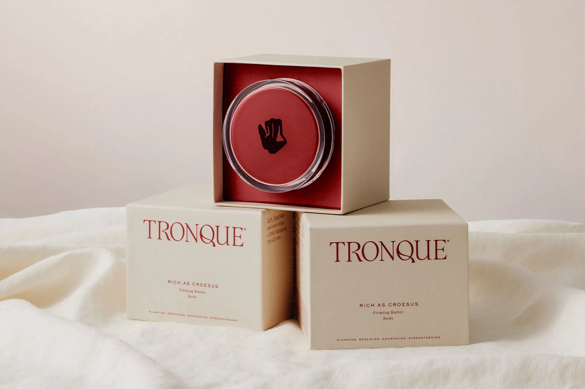 TRONQUE skincare product displayed in box on top of two other boxes against a cream-colored draped fabric backdrop. Beauty shot.