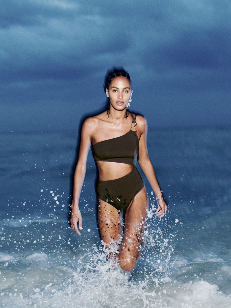 A woman in a one-shoulder swimsuit walks through the ocean water, with a dark blue sky visible in the background on a sunny day.