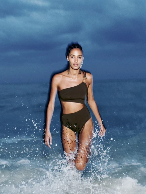 A woman in a one-shoulder swimsuit walks through the ocean water, with a dark blue sky visible in the background on a sunny day.