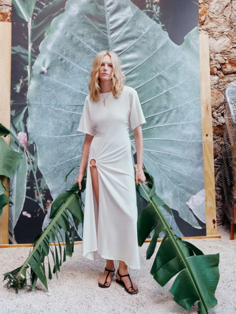 A fair-skinned adult woman poses in a white dress with a thigh slit, holding green banana leaves indoors against a backdrop of a large green leaf.