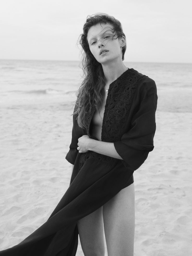 Black and white portrait of a female model posing on a beach wearing an open robe, with ocean and sky visible in the background.