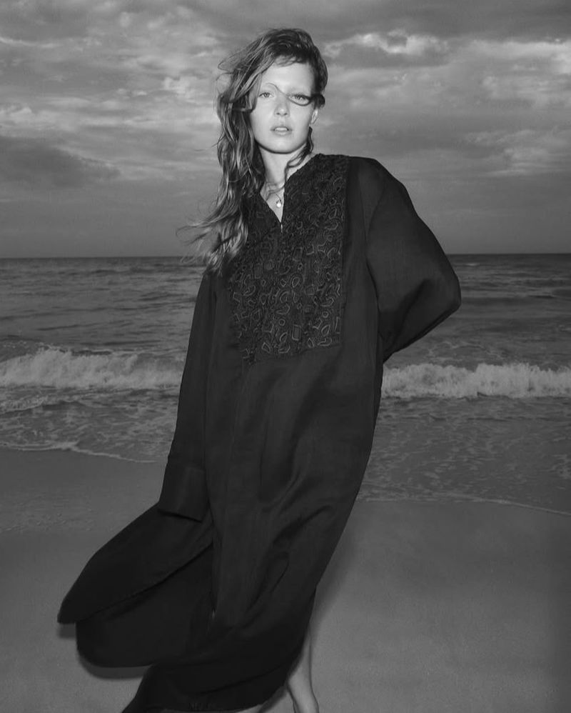 Black-and-white portrait of woman in embroidered black dress standing on a beach with a cloudy sky and ocean backdrop at dusk.