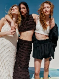 Three female models posing outdoors with arms around each other against a bright blue sky and sandy beach background.