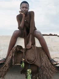 Model wearing a brown dress seated on a concrete ledge holding a large fringed brown bag against an overcast sky.