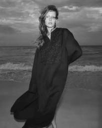 Monochrome fashion photograph of a model in a dark embroidered kaftan standing on a sandy beach with ocean waves behind.