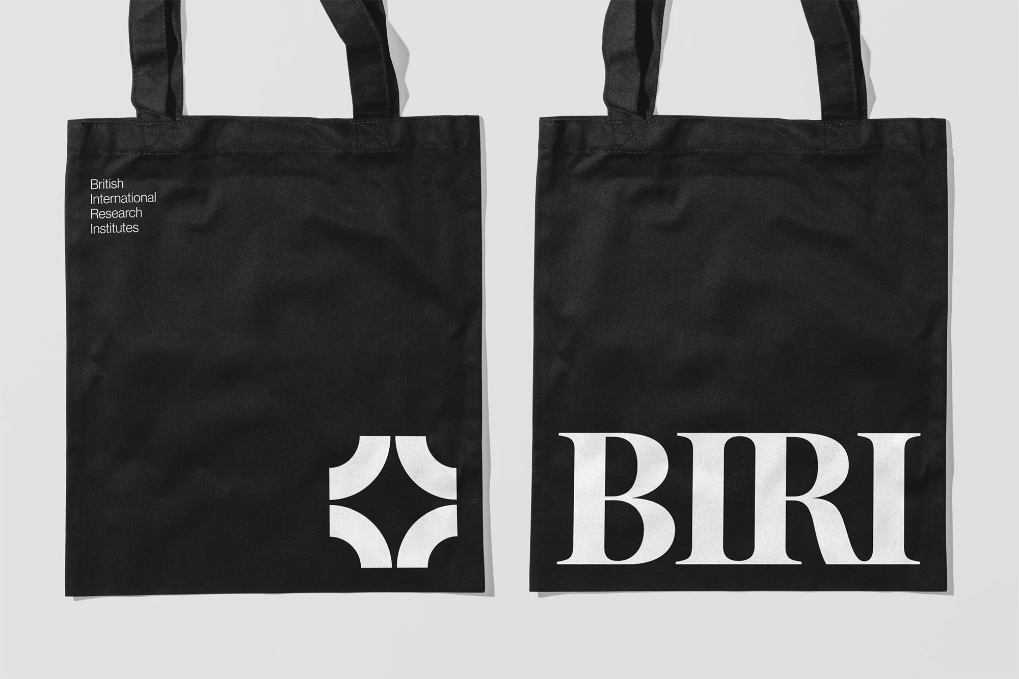 Two black tote bags on a white background, each featuring a white logo and text: 'British International Research Institutes' and 'BIRI'.