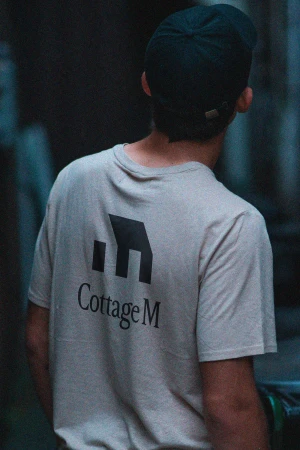 Back view of a person in a T-shirt with the 'Cottage M' logo and a cap, set against a muted, dark background, focusing on branding.