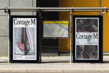 Two vertical advertising posters for Cottage M displayed in a glass shelter on a city sidewalk.