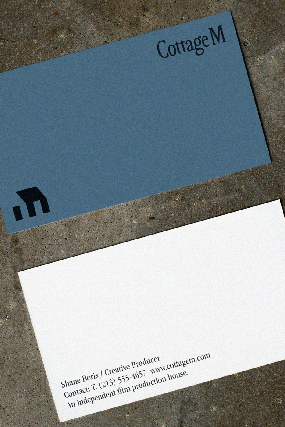 Business card for Cottage M featuring a slate blue front with a black logo and a white reverse side with contact details on stone.