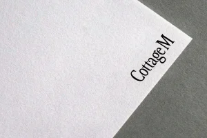 Close-up of textured white paper card featuring the black CottageM logo placed on a grey surface.