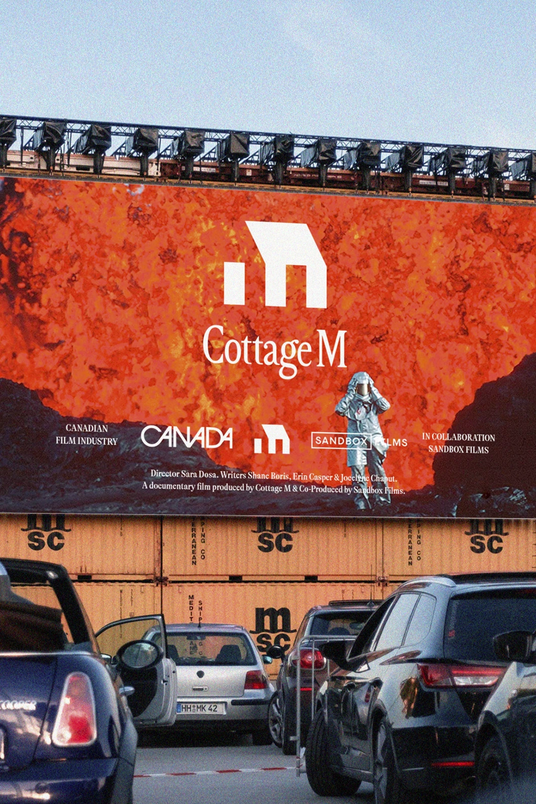 Large red promotional billboard displaying the Cottage M logo mounted above stacked shipping containers and cars parked on a street