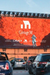 Large red promotional billboard displaying the Cottage M logo mounted above stacked shipping containers and cars parked on a street