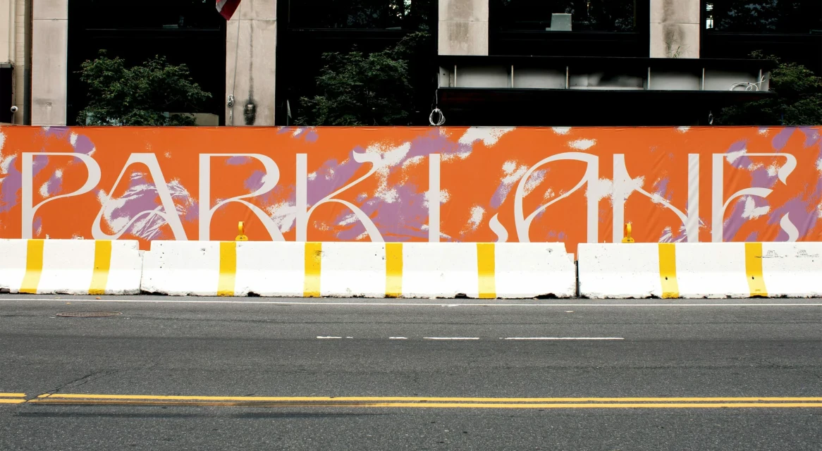 Urban scene featuring street-level view of asphalt road, concrete barriers with yellow stripes, and a large orange sign that reads PARKLANE.
