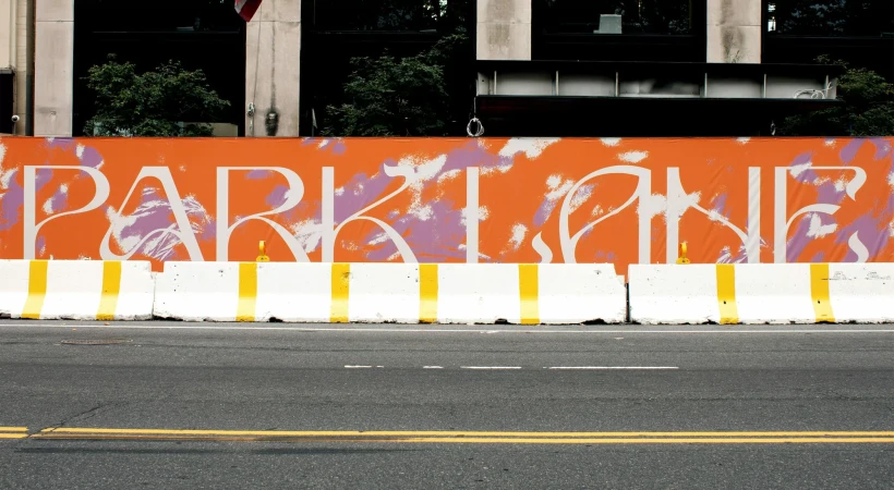 Urban scene featuring street-level view of asphalt road, concrete barriers with yellow stripes, and a large orange sign that reads PARKLANE.