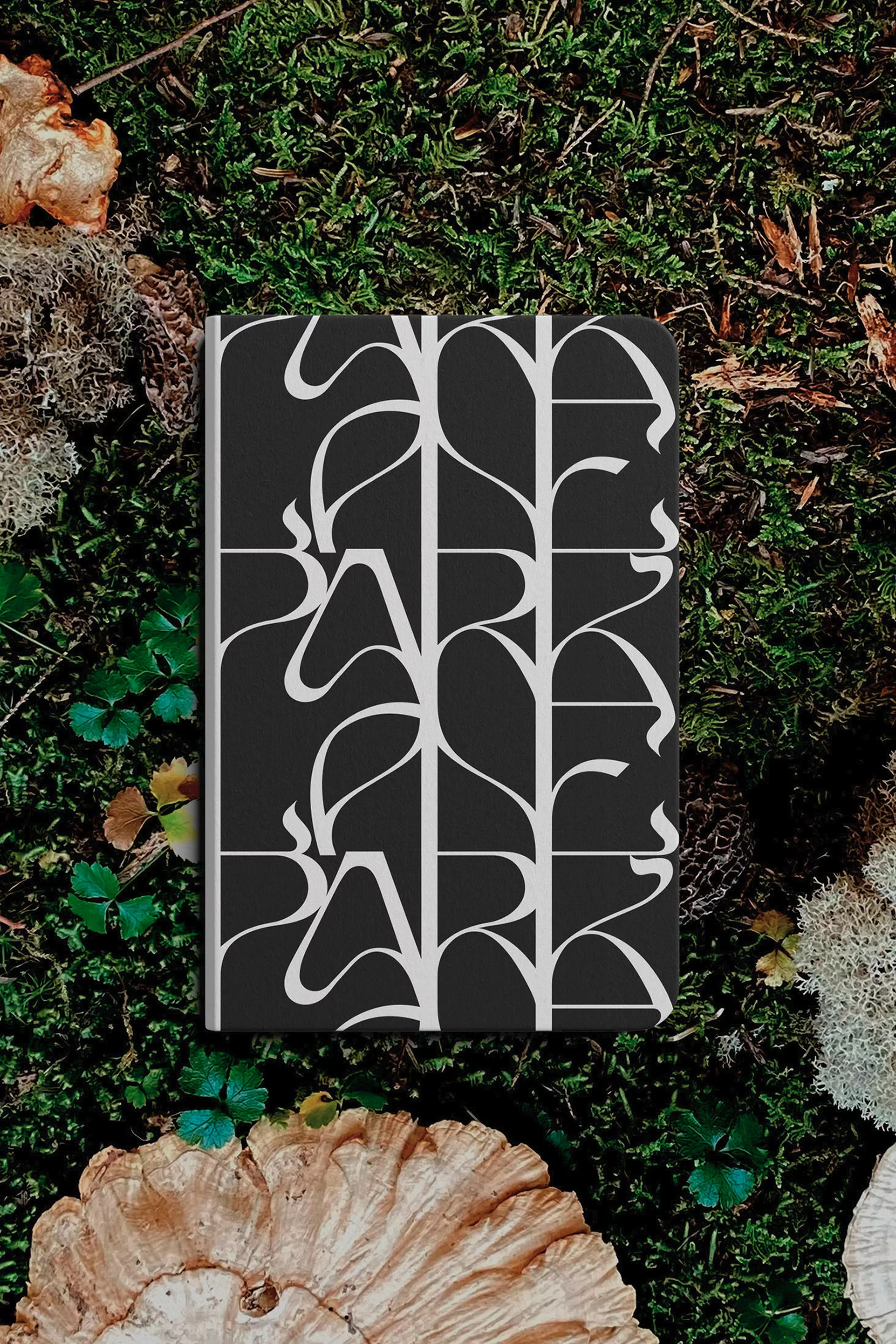 Black and white patterned book lying on green moss, surrounded by mushrooms and leaves in a natural outdoor setting.