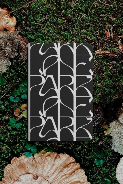 Black and white patterned book lying on green moss, surrounded by mushrooms and leaves in a natural outdoor setting.