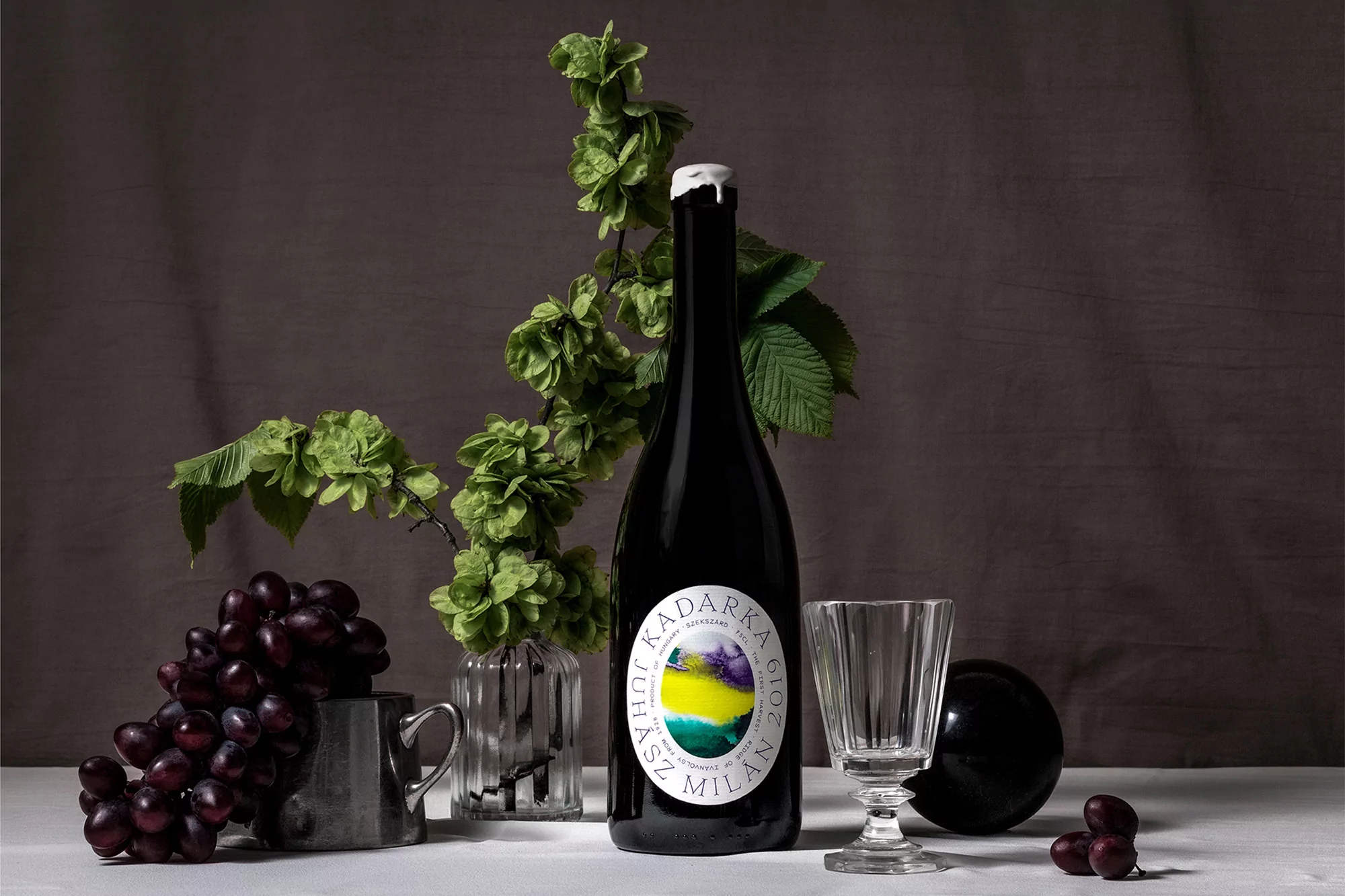 Still life shot of a dark bottle with a white label, glass cup, grapes, and green hops in a studio setting, against a grey background.
