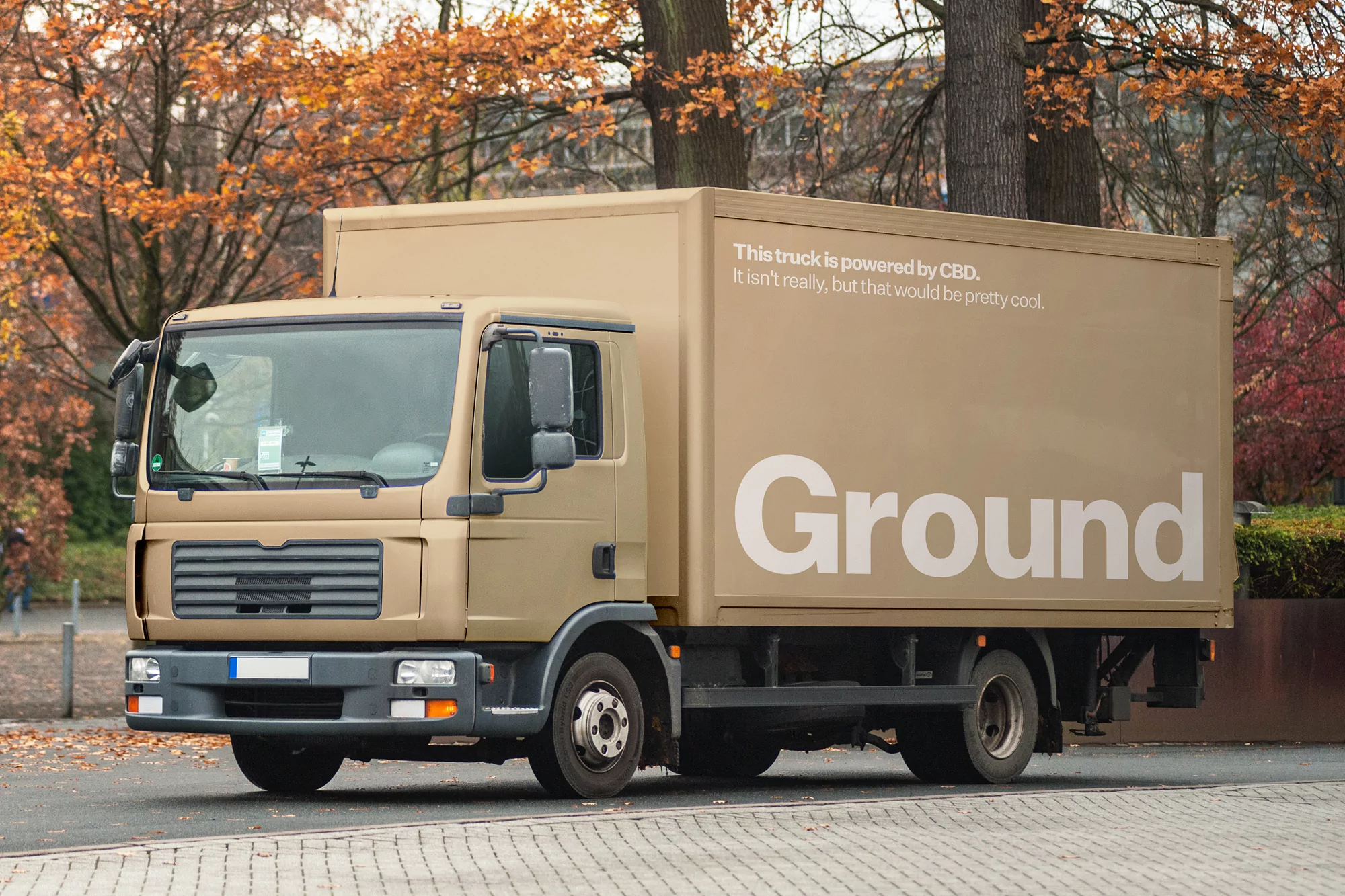 A tan-colored delivery truck with the word 'Ground' displayed on its side parked on a paved road with fall trees in the background.