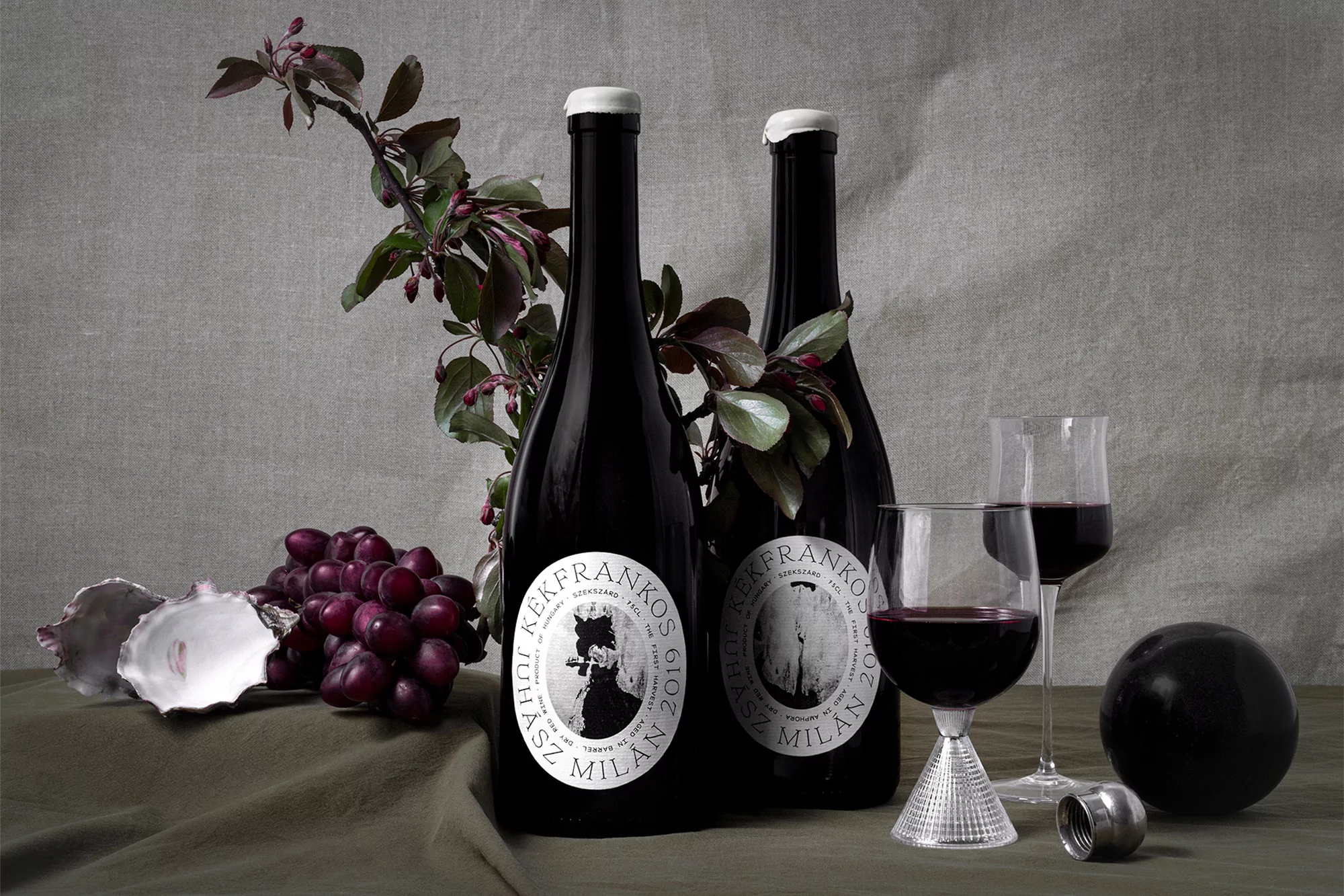 Two bottles of dark red wine sit with two glasses, red grapes, oyster shell, and a black sphere on a draped olive cloth.
