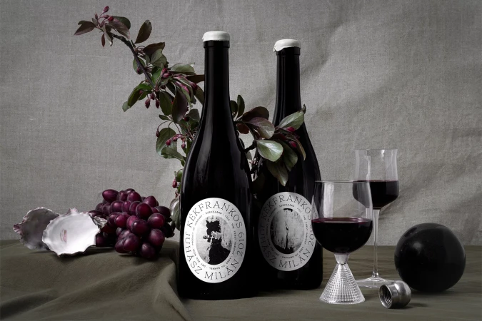 Two bottles of dark red wine sit with two glasses, red grapes, oyster shell, and a black sphere on a draped olive cloth.