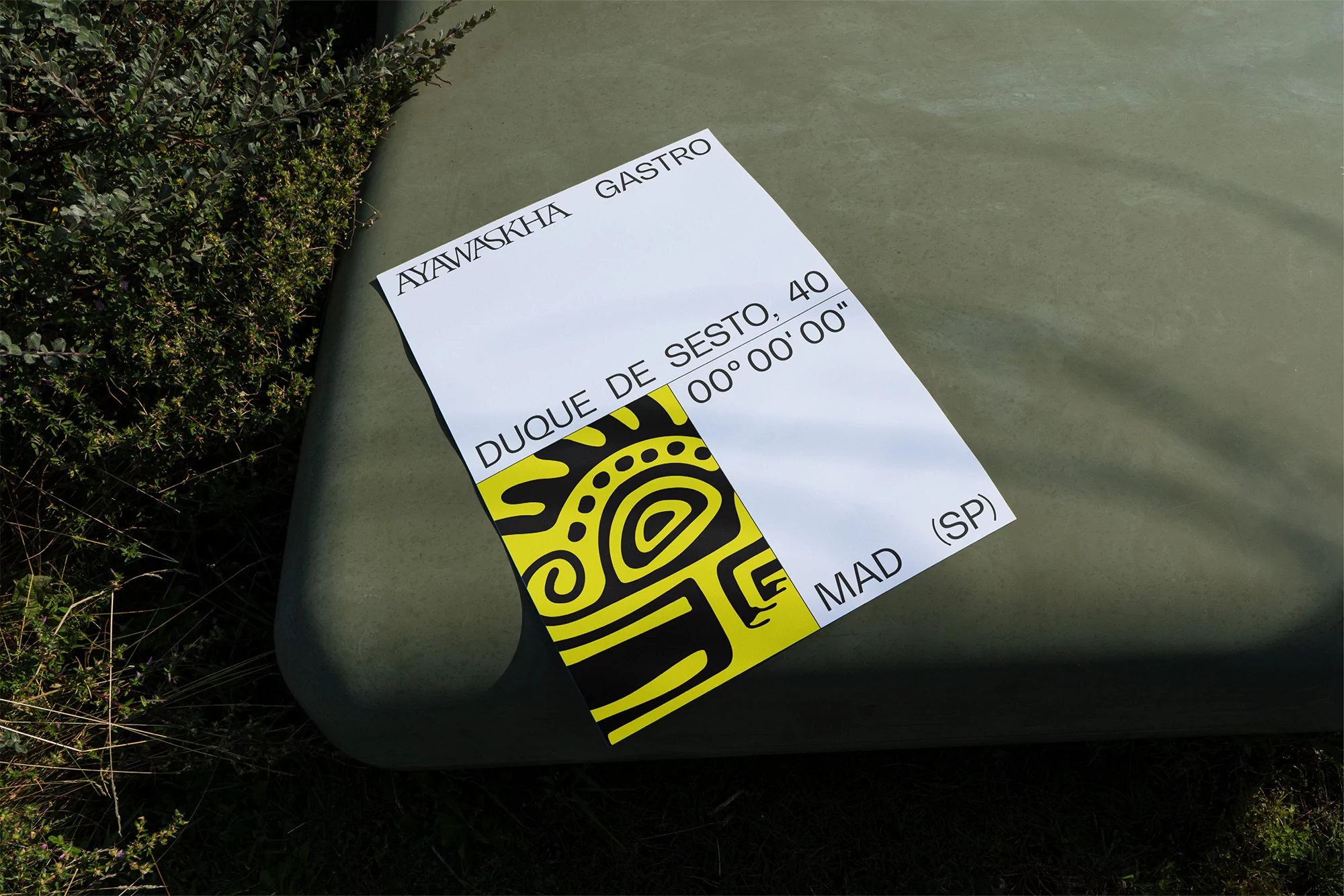 Promotional flyer rests on an olive-green surface outdoors, advertising Ayawaskha Gastro at Duque de Sesto, 40 in Madrid, Spain.