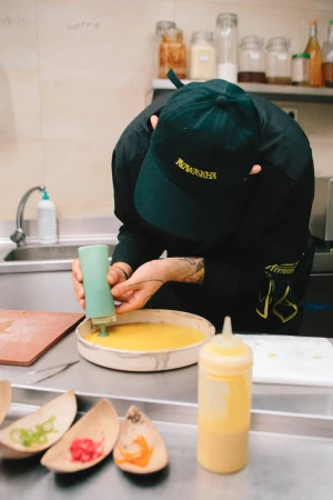 Chef in black uniform carefully decorates a yellow sauce on a plate in a commercial kitchen, using a green squeeze bottle.