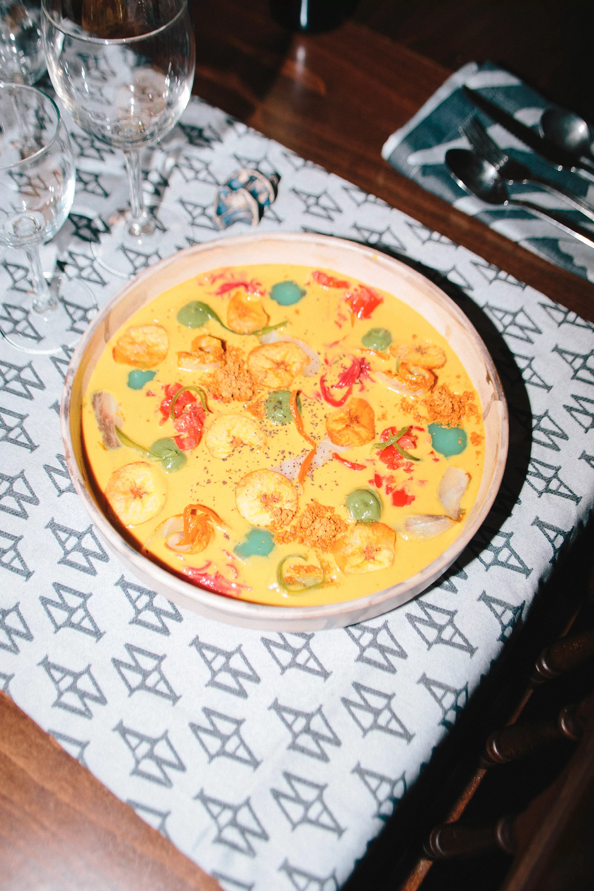Overhead shot of a wooden bowl filled with a creamy yellow dish topped with sliced bananas, tomatoes, and herbs on a patterned tablecloth.