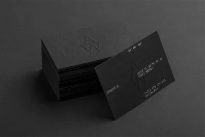 A stack of dark business cards with debossed geometric monogram logos and minimal typography resting on a dark textured surface.