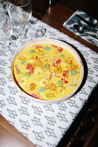 Overhead view of a yellow culinary dish in a wooden bowl served on a patterned tablecloth with wine glasses and cutlery.