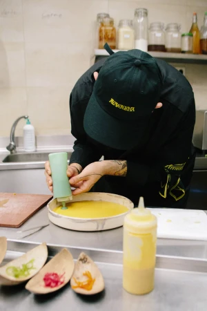 A chef in a black uniform and cap pouring sauce from a squeeze bottle onto a dish in a commercial kitchen.