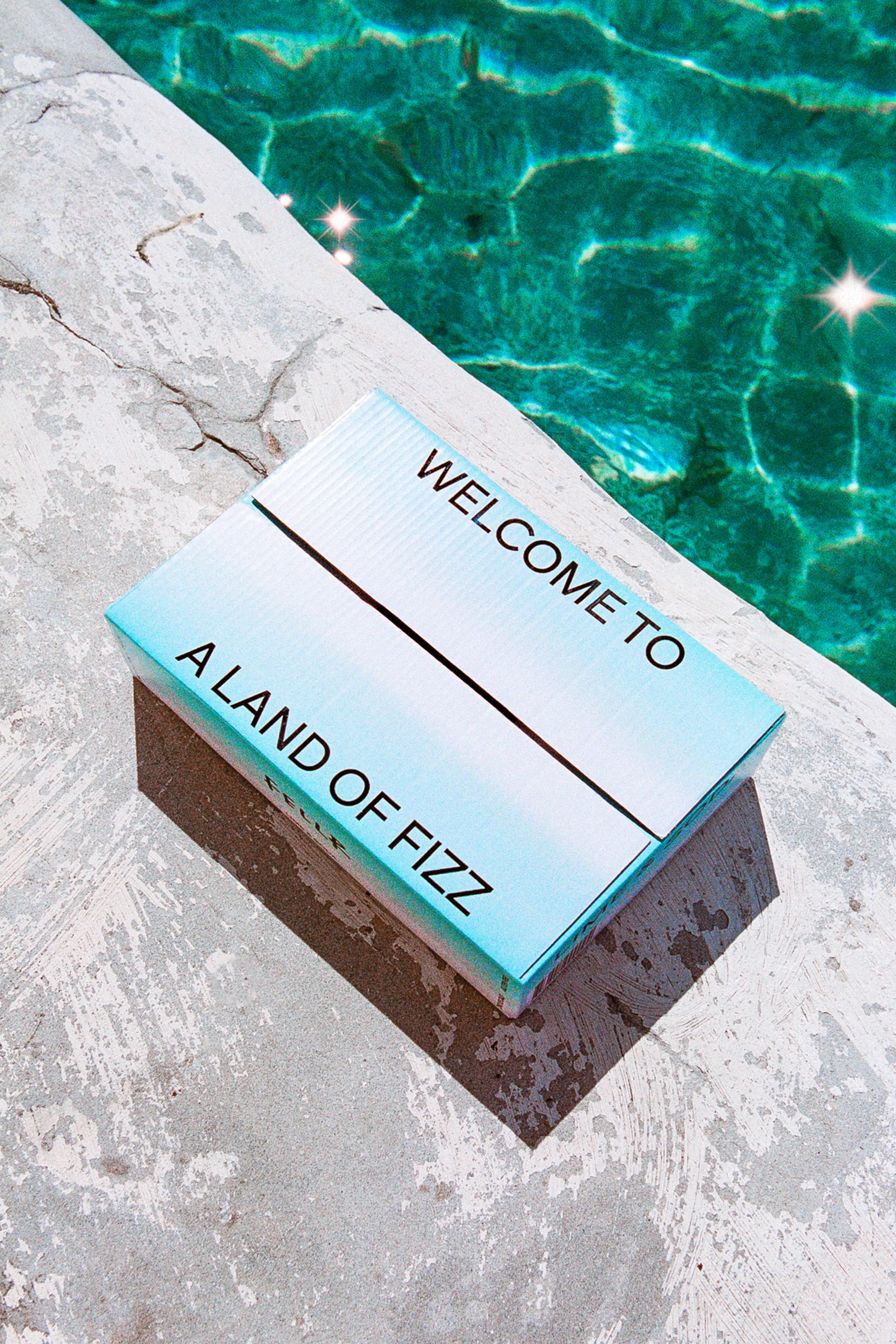 Sky-blue and white box sitting on a concrete surface next to pool water with a message 'Welcome to A Land of Fizz'.