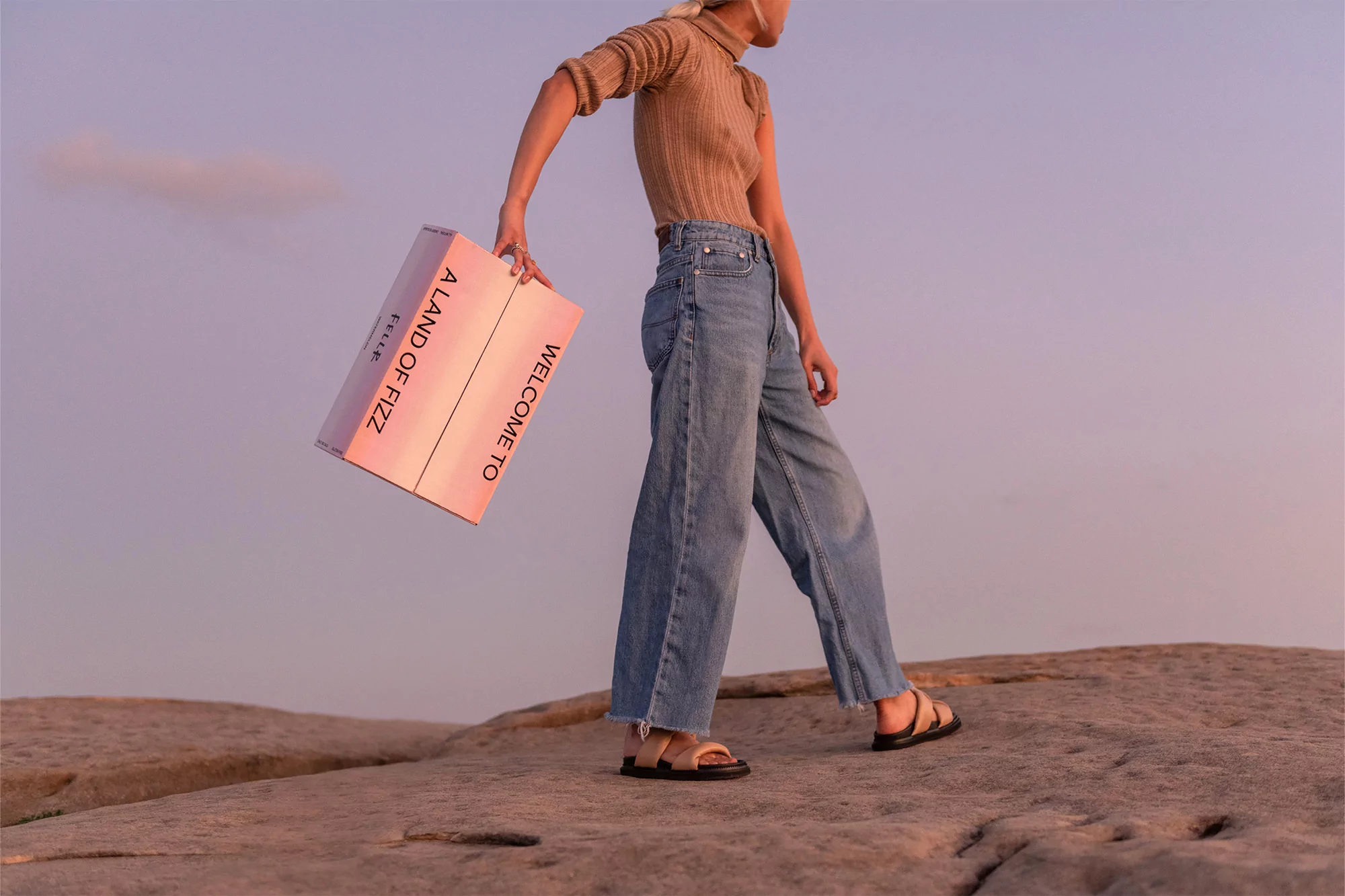 A female model in a turtleneck sweater and wide-leg jeans poses on a rock formation while holding a branded box in a desert setting.