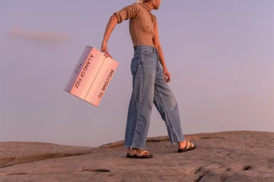 Model standing on rocky terrain carrying a pink branded box that reads Welcome To A Land Of Fizz for FELLR.