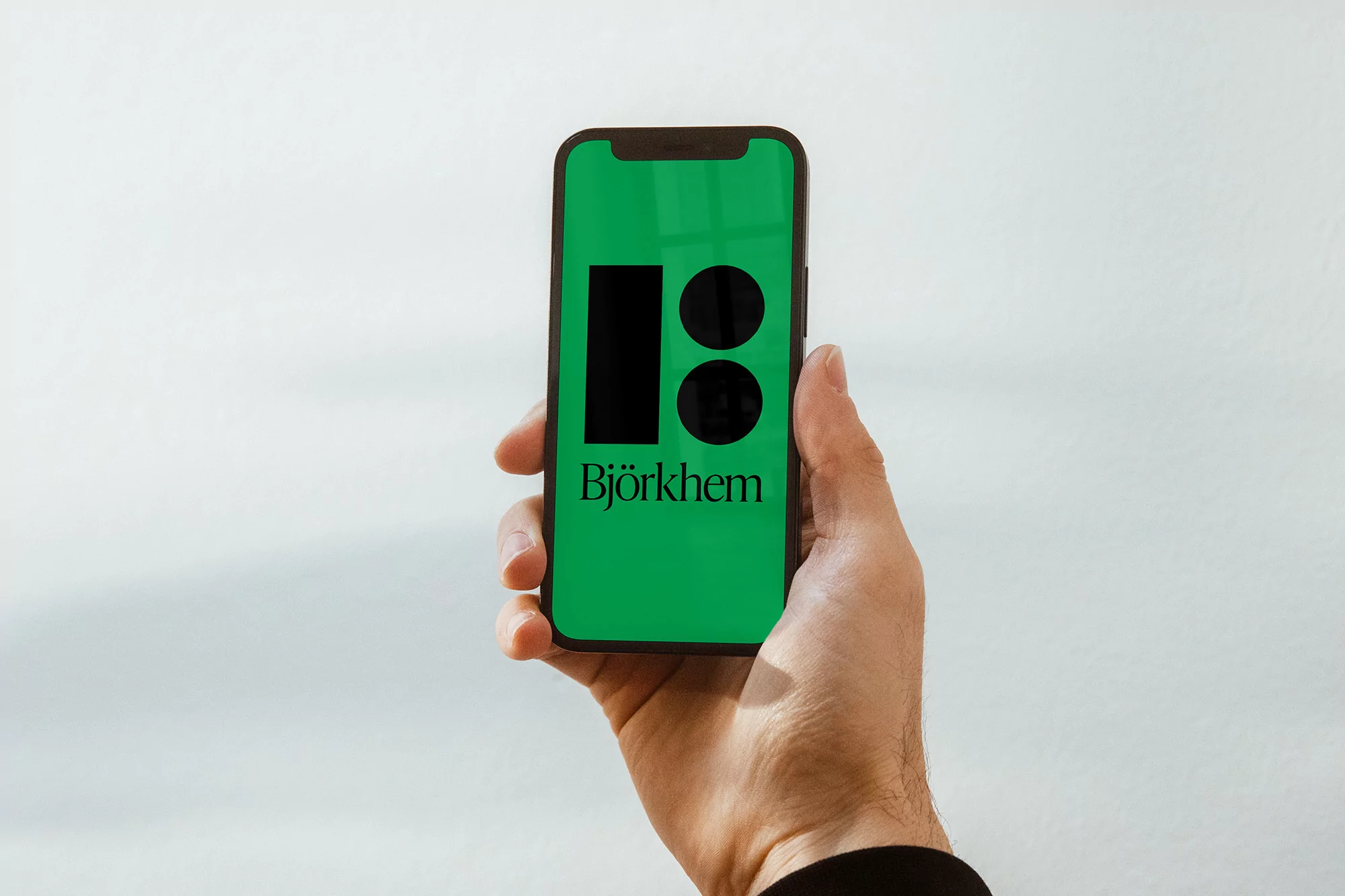 A hand holds a smartphone displaying a green screen with a logo of a black rectangle and two black circles with the text 'Björkhem'.