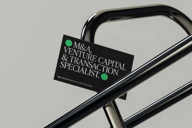 Black business card with white typography and green dots tucked into a metal railing against a grey background.