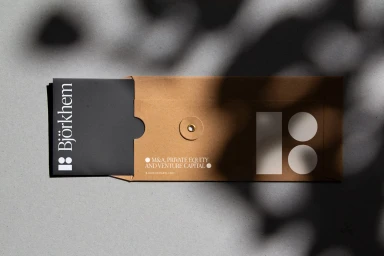 Kraft paper envelope and black folder with Bjorkhem branding resting on a textured grey concrete surface under dappled shade.