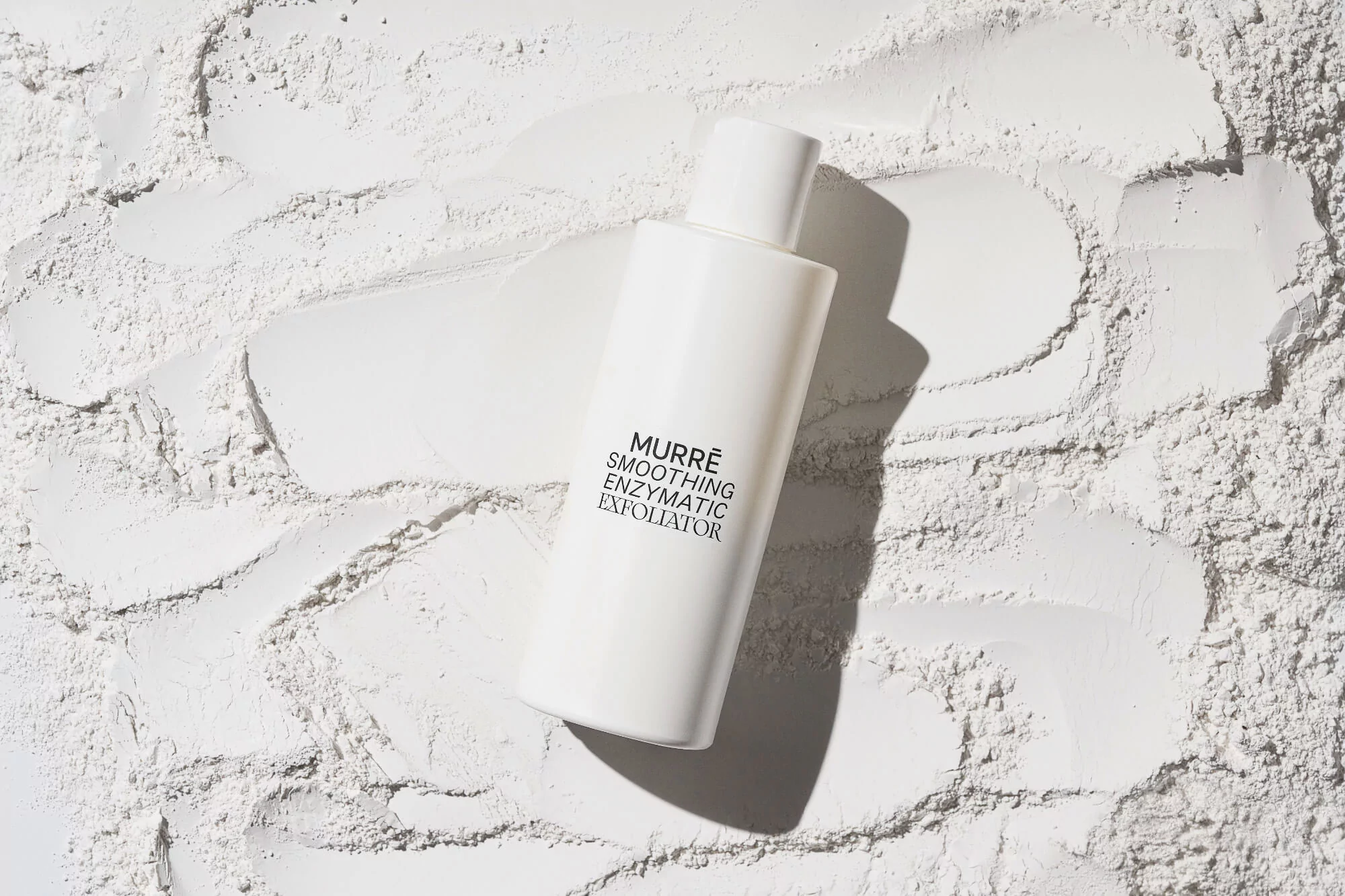 White bottle of Murre smoothing enzymatic exfoliator sits in a bed of white powder under bright studio lighting. Beauty product shot.