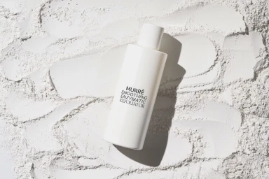 White skincare bottle with black text resting on textured white powder surface in harsh studio lighting