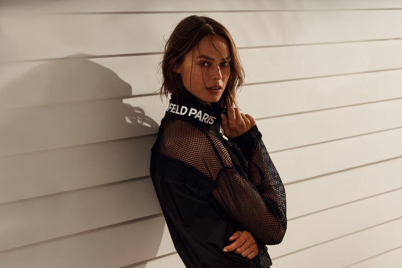 A woman poses indoors wearing a black mesh jacket with 'KARL FELD PARIS' printed on the collar, with a neutral, minimalist background.