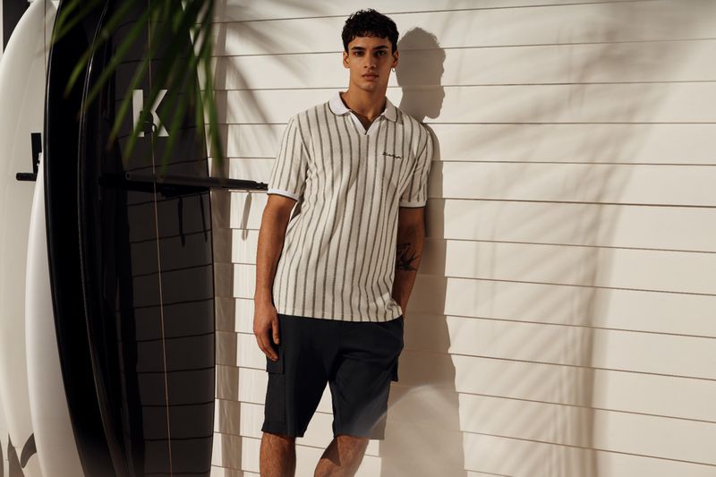 Handsome man in a striped polo shirt and black shorts poses against a white wall with surfboards and palm leaf shadows.